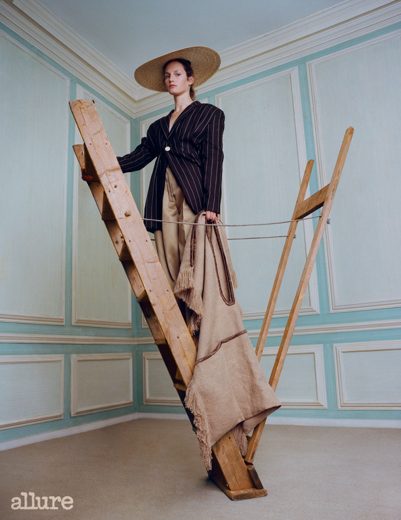 Feature, accessories, classic, heirloom, neutral palette, model, curly hair, natural look, standing on upside-down ladder, holds oversized jute bag, wears striped wool blazer, straw hat, beige trousers